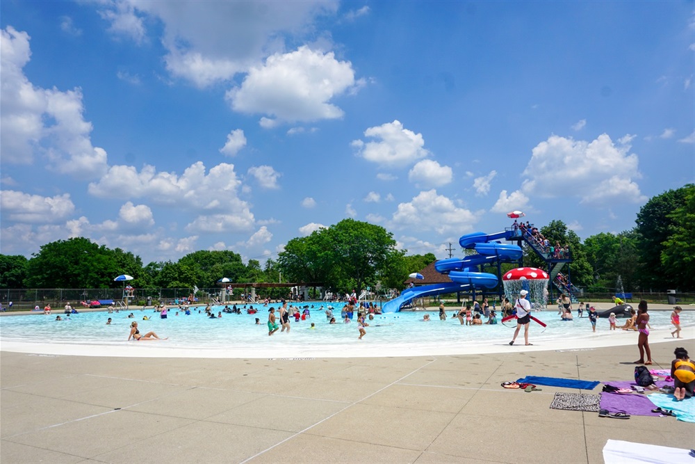 People swim in a large, zero-depth entry pool with a spray feature and water slide.