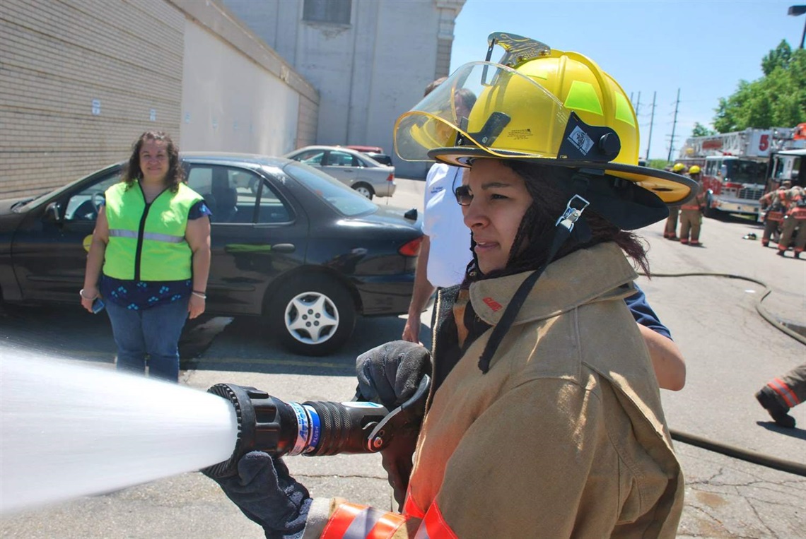 Young woman of color holding fire hose and spraying water during the Fire Fighter for a Day event.
